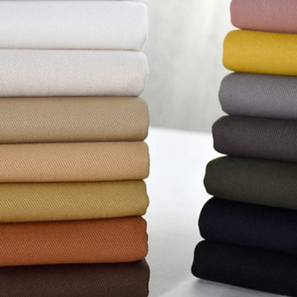 A neatly stacked collection of fabric rolls, featuring two rows of twill cotton fabric in various colors, with a focus on beige, white, yellow, gray, black, and darker shades.