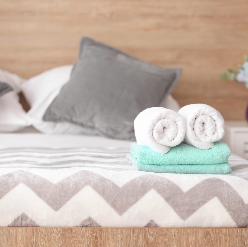 Stacked clean white towels, with two rolled towels on top, placed on a bed with a grey and white zigzag-patterned bedsheet. In the background, there is a grey pillow against a wooden headboard.