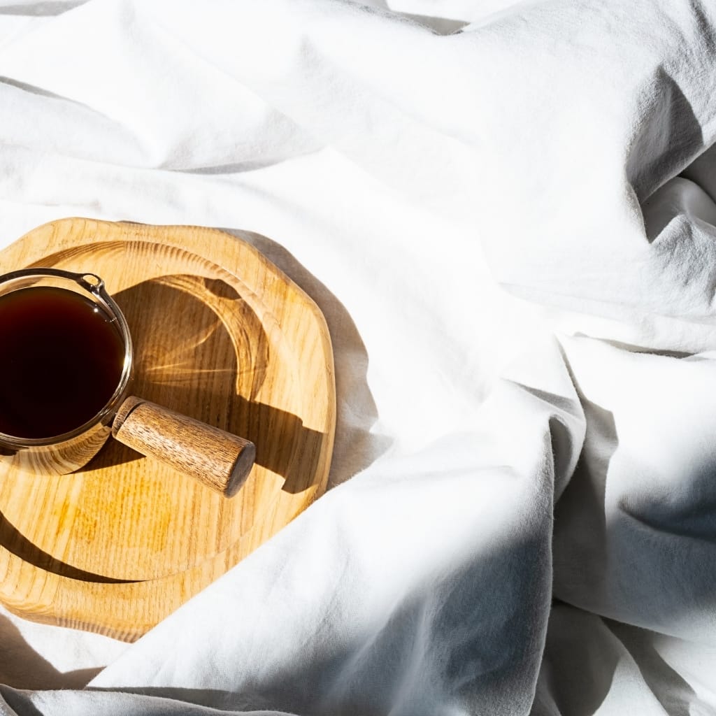 A glass cup of dark coffee placed on a round wooden tray, set on a white, crumpled bedsheet. The sunlight casts a shadow of the cup and tray on the sheet.