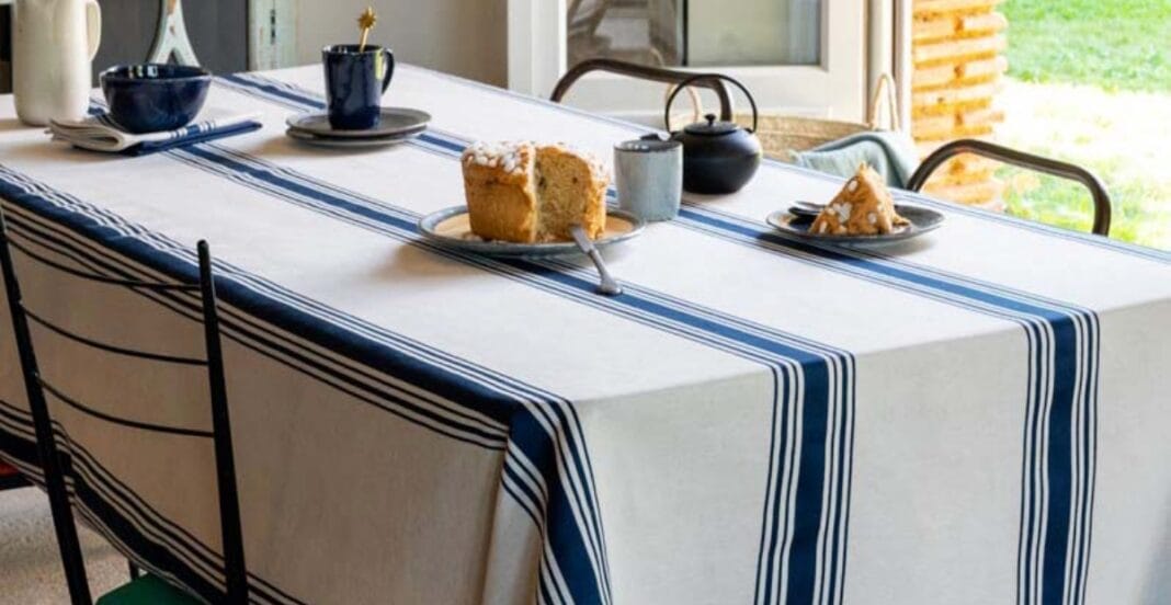 : A dining table covered with a white tablecloth featuring blue and navy stripes. The table has slices of cake on plates, a teapot, a cup, and some bowls, set near a window with a bright outdoor view.