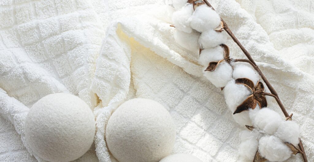 Two white bath bombs are placed on a soft, textured white towel. A branch of cotton with fluffy white cotton balls and brown dried calyxes is laid across the towel next to the bath bombs.