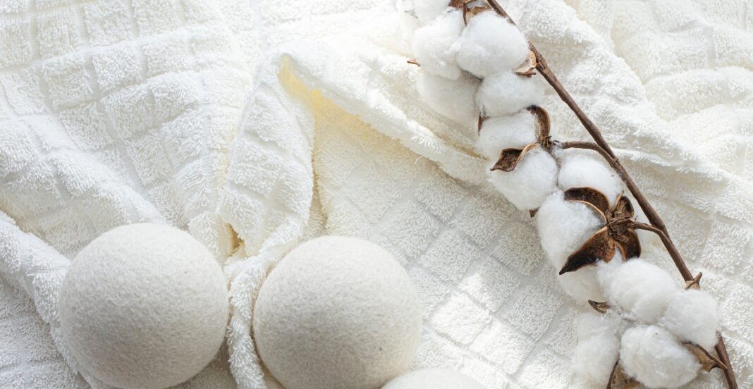 Two white bath bombs are placed on a soft, textured white towel. A branch of cotton with fluffy white cotton balls and brown dried calyxes is laid across the towel next to the bath bombs.