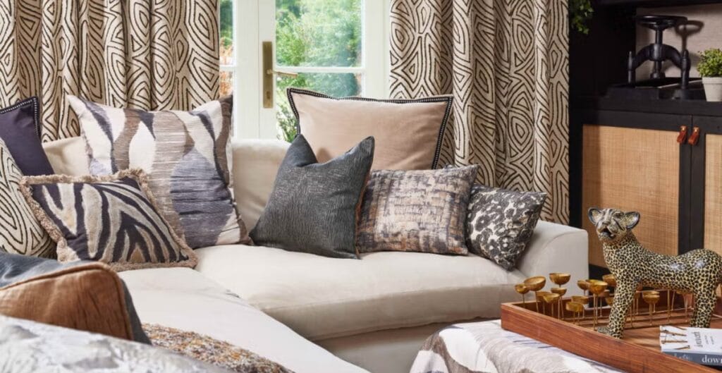 A cozy living room featuring a white couch adorned with numerous decorative pillows in various patterns and textures. Behind the couch, there are patterned curtains and a window showing greenery outside. On the right side, there is a black and wood cabinet with small potted plants on top. In the foreground, there is a wooden tray with a decorative leopard figurine and other objects.