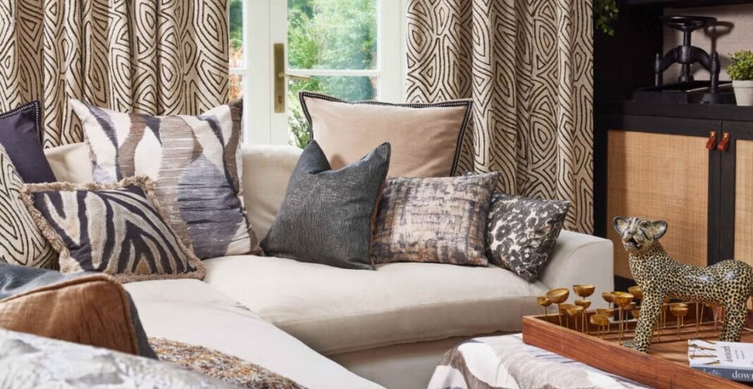 A cozy living room featuring a white couch adorned with numerous decorative pillows in various patterns and textures. Behind the couch, there are patterned curtains and a window showing greenery outside. On the right side, there is a black and wood cabinet with small potted plants on top. In the foreground, there is a wooden tray with a decorative leopard figurine and other objects.