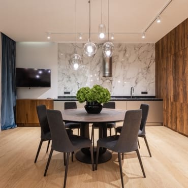 A modern kitchen and dining area with a round black table, six upholstered chairs, a marble backsplash, pendant lighting, and wooden accents.