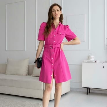 A young woman in a vibrant pink belted shirt dress with short sleeves, standing inside a modern, minimalistic room with white walls and a beige sofa.