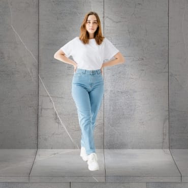 A young woman standing against a concrete wall, wearing a white polyester T-shirt, distressed denim jeans, and white sneakers.