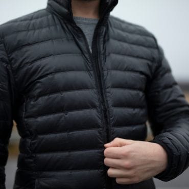 A person wearing a black, quilted weather-resistant polyester jacket with a high collar, zipped up, with their hand gripping the zipper.
