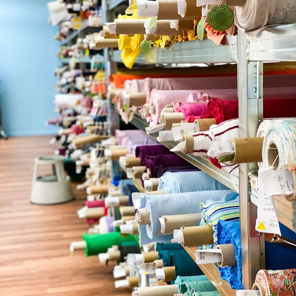 A store display with multiple rows of colorful fabric rolls, primarily cotton material, arranged on shelves.