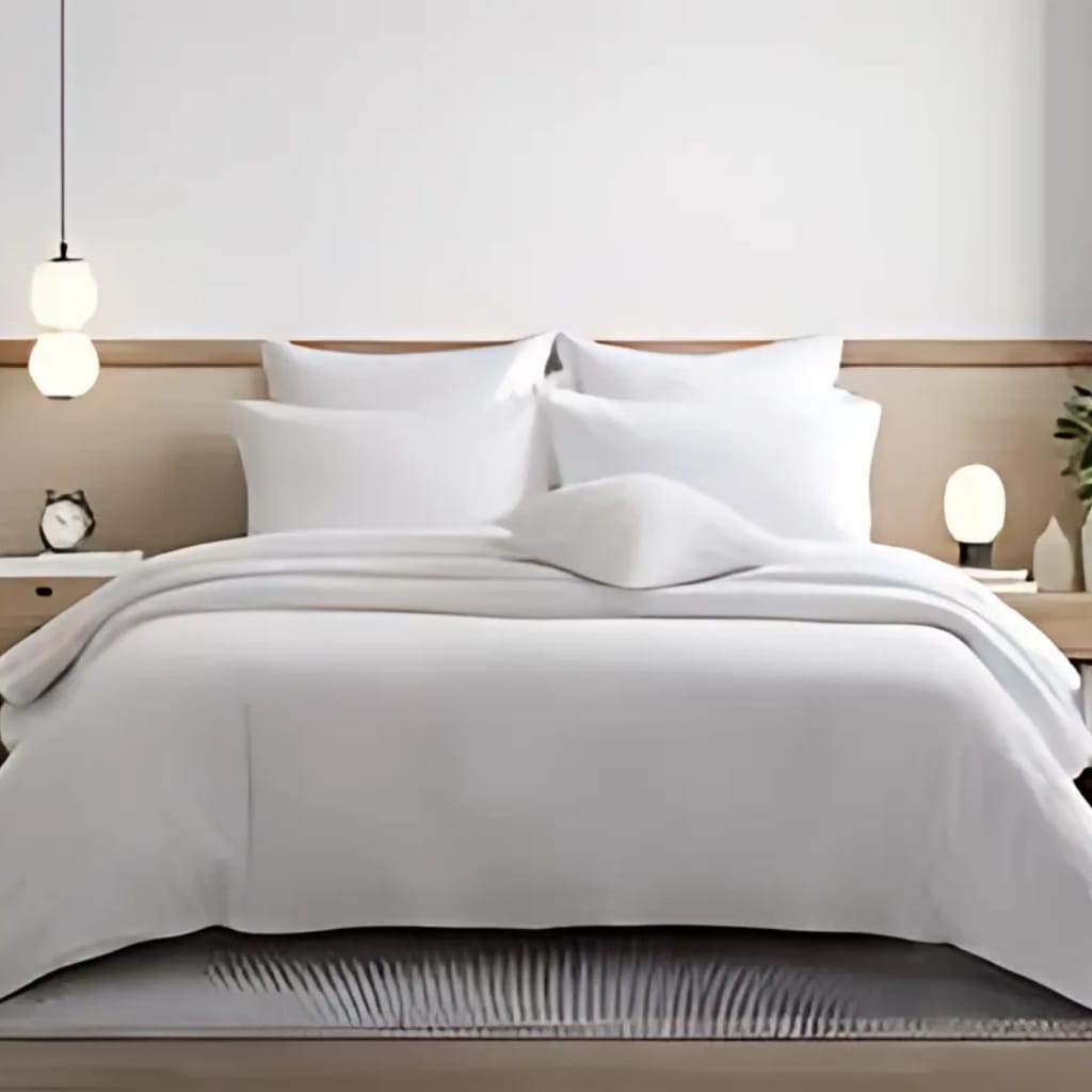 A neatly made bed with white bedding, multiple white pillows, and a simple wooden headboard. There are two small bedside tables, each with a lamp, and a small decorative plant on the right side.