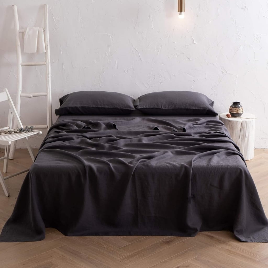 A neatly made bed with dark grey bedding and pillows in a minimalist bedroom. On the left side, there's a white wooden chair with some jewelry draped over it, and on the right, a small white bedside table with decorative objects. The background features a plain white textured wall, and a modern light fixture hangs above the bed.