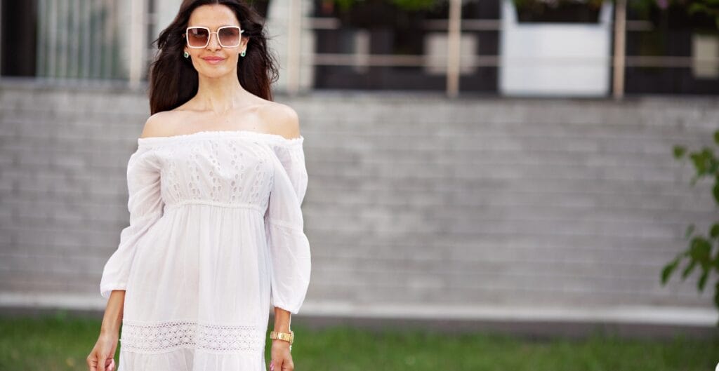 A woman standing outdoors in front of a modern building with gray brick walls. She is wearing large sunglasses, a white off-shoulder dress with lace details, and has long wavy dark hair.
