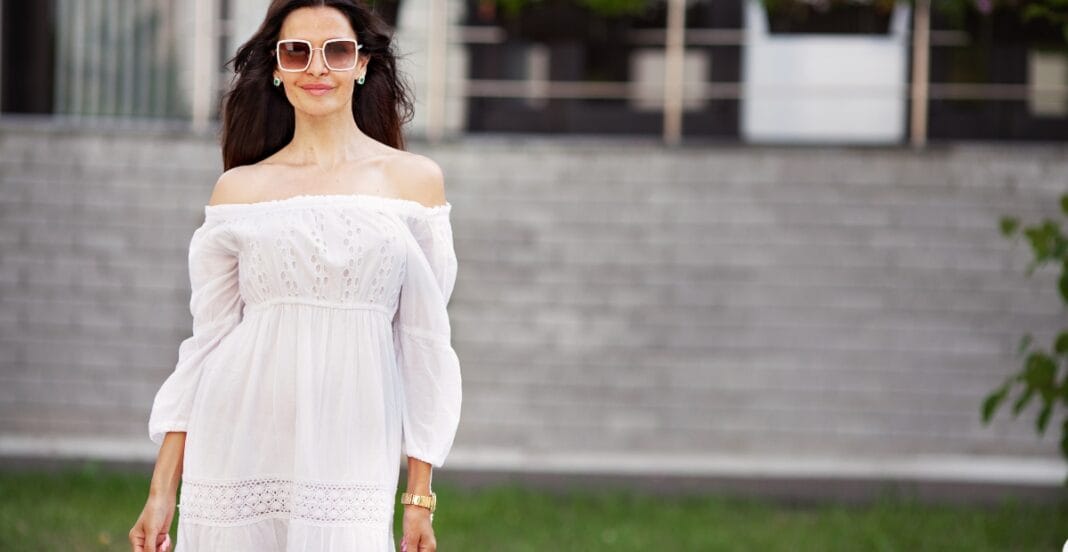 A woman standing outdoors in front of a modern building with gray brick walls. She is wearing large sunglasses, a white off-shoulder dress with lace details, and has long wavy dark hair.