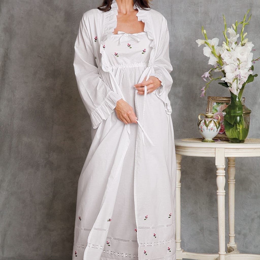 A woman dressed in white, wearing a long, embroidered, and ruffled dress, standing next to a small table with a vase of white flowers and a decorative picture frame on it.