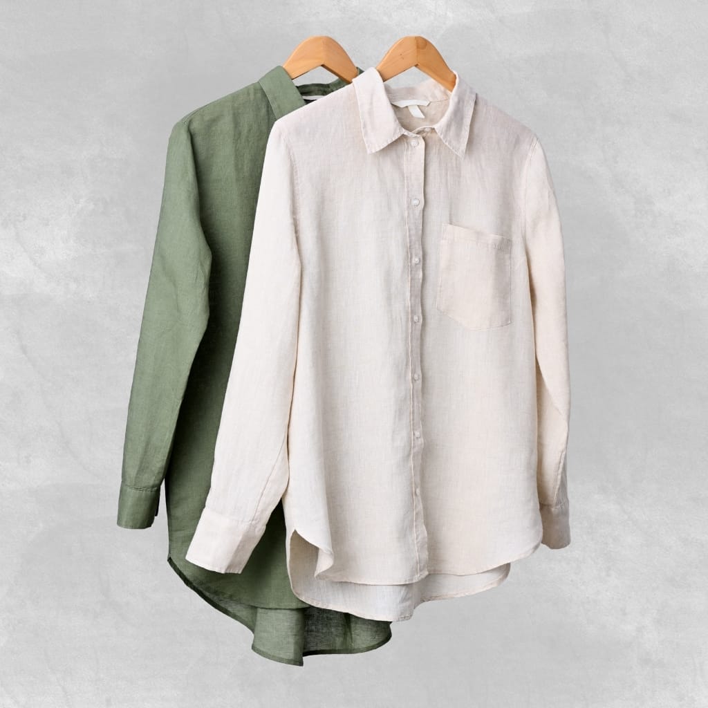 Two long-sleeve linen shirts, one in green and the other in off-white, hanging on wooden hangers against a light gray background.