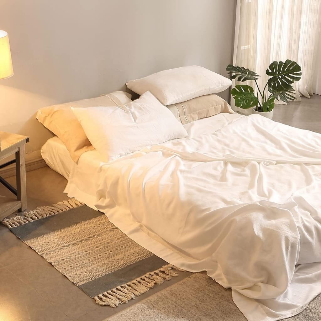 A neatly made bed with white bedding and multiple pillows, positioned beside a nightstand with a lamp and a potted monstera plant, in a minimalistic and bright bedroom.