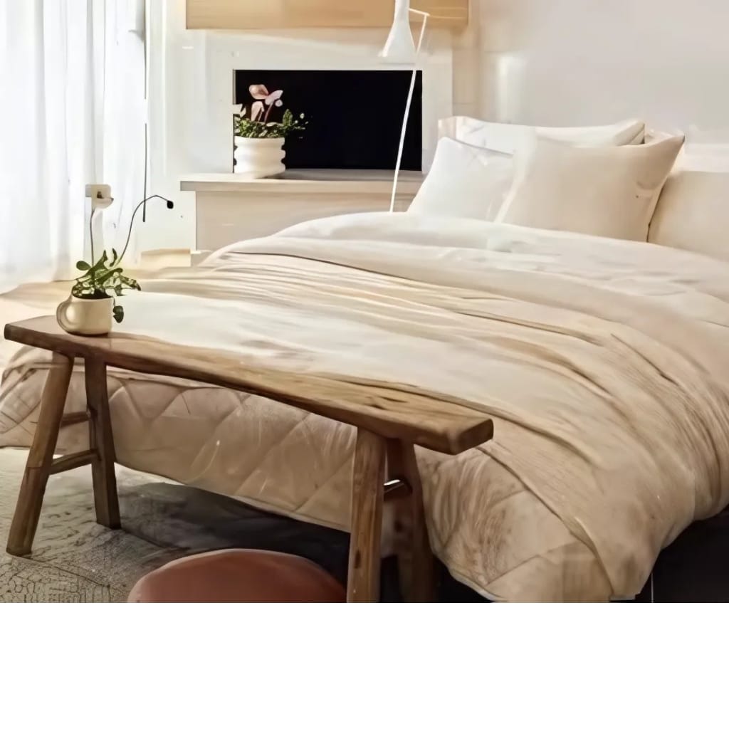 A cozy bedroom featuring a neatly made bed with white linens, a wooden bench at the foot of the bed holding a small potted plant, and a window with sheer white curtains letting in soft light. There is a TV on the wall across from the bed, and a side table with a stylized lamp.