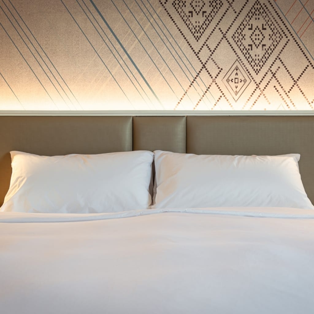 A neatly made bed with white linens and pillows, set against a modern headboard and a decorative wall featuring geometric patterns and lines with soft lighting.