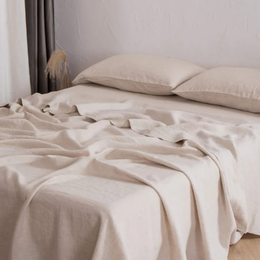A neatly made bed with cream-colored bedsheets and two matching pillows on a white wall background. There are some folds and slight wrinkles on the bedsheets, giving a cozy and inviting appearance.