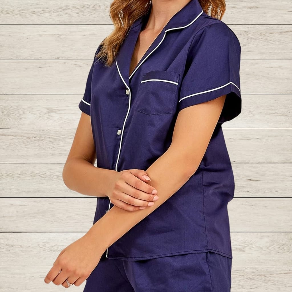 A person wearing a navy blue satin pajama set with white piping, standing against a light wooden background. The person is shown from the shoulders down, with their arms loosely crossed in front of them.