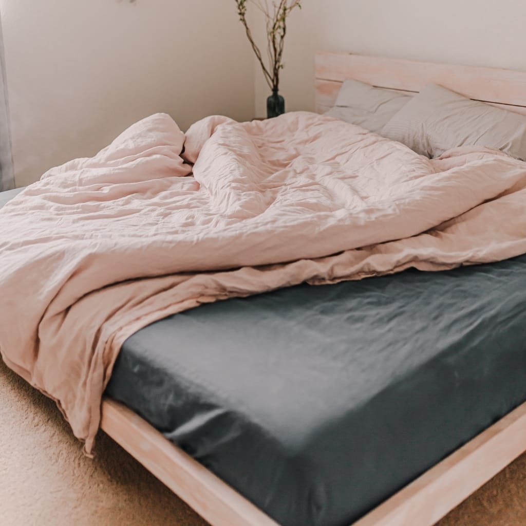 A neatly made bed with a dark grey fitted sheet and a soft, pale pink duvet cover. The duvet is slightly rumpled, and two pillows with beige pillowcases rest against a light wooden headboard. A small vase with a dried branch decorates the corner of the room.