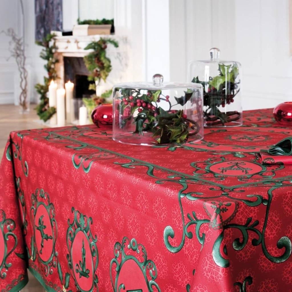A decorated holiday table with a red tablecloth featuring green ornate patterns. There are glass domes containing holly and berries, and a small red ornament on the table.
