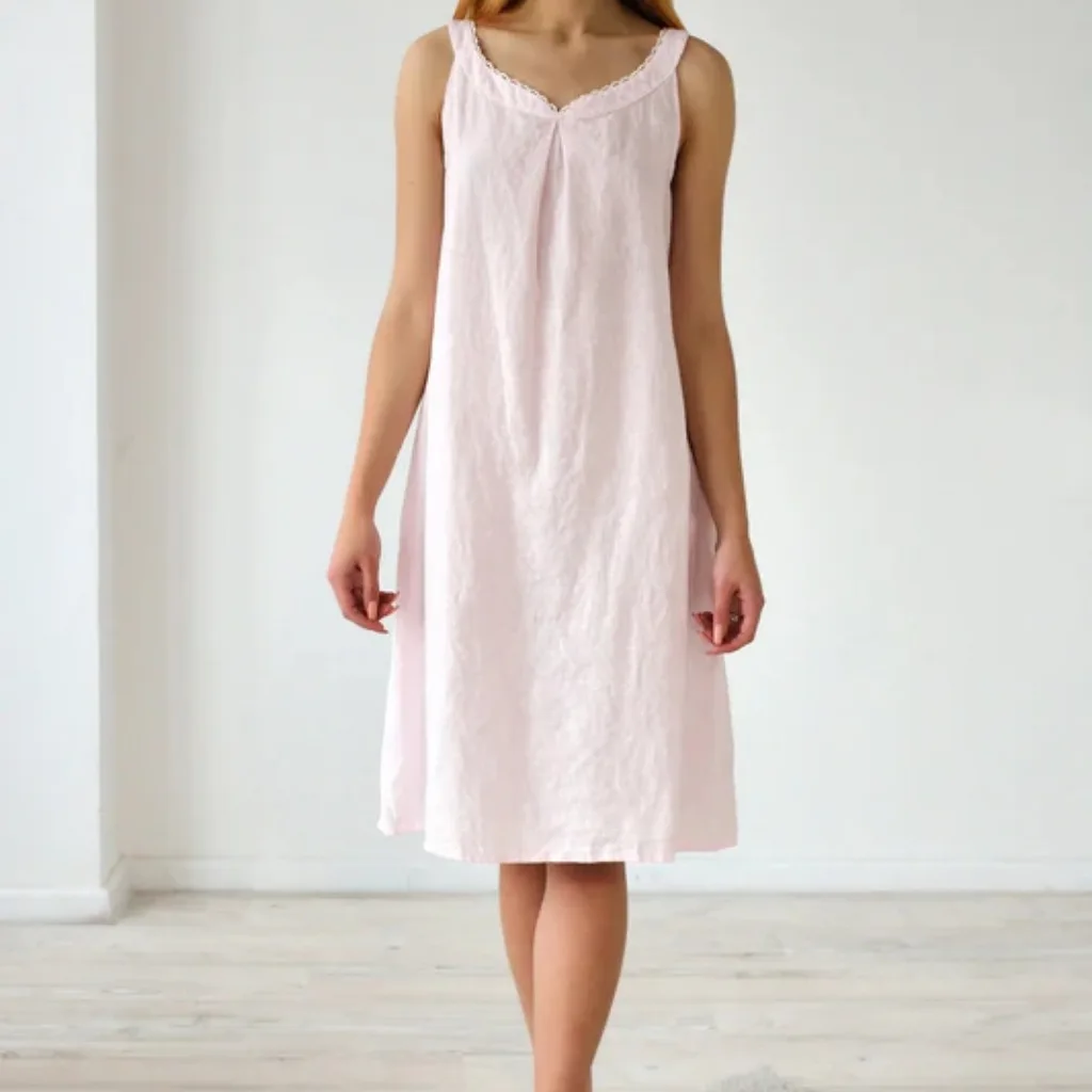 A woman wearing a light pink, sleeveless, knee-length nightgown with lace trim around the neckline, standing in a minimalistic room with light-colored wooden flooring and plain white walls.