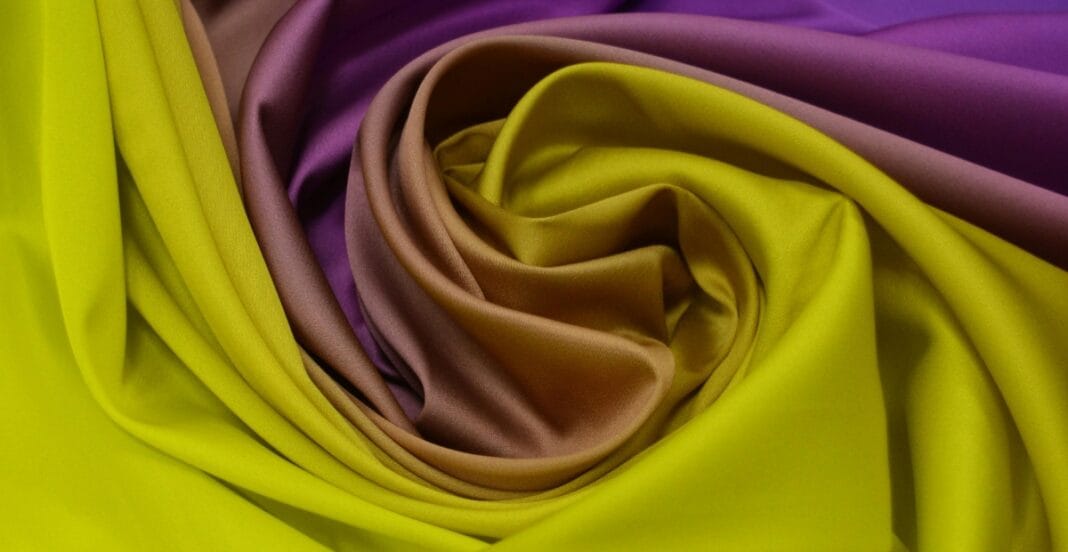 A close-up of colorful, shiny polyester fabric in shades of yellow, pink, and purple, arranged in a spiral pattern.
