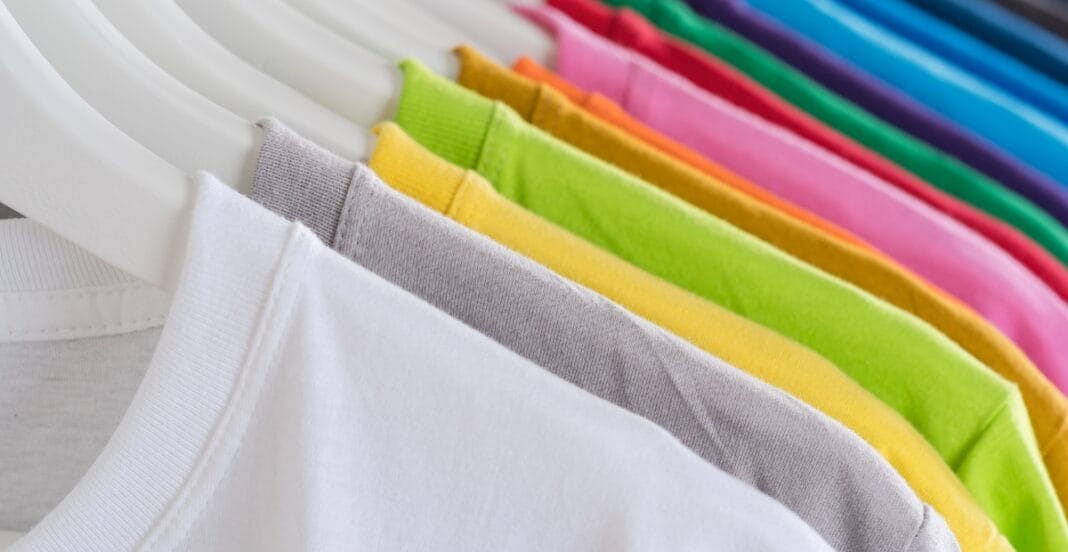 A row of colorful cotton t-shirts hanging on white hangers, showcasing various shades including white, gray, yellow, green, pink, purple, and blue.