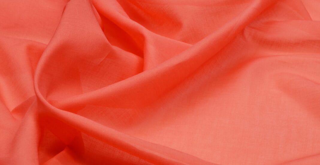 A close-up image of coral-colored cotton batiste fabric with gentle folds, highlighting its smooth and lightweight texture.