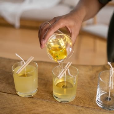 A person pouring liquid polyester resin into two clear glasses, each with a straw, on a wooden surface.