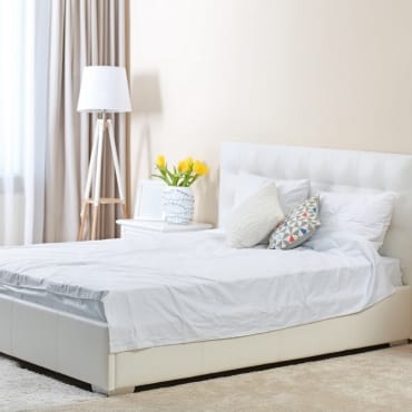 A cozy bedroom featuring a white bed with white bedding, decorative pillows, a side table with a vase of yellow flowers, a modern white floor lamp, and beige curtains.