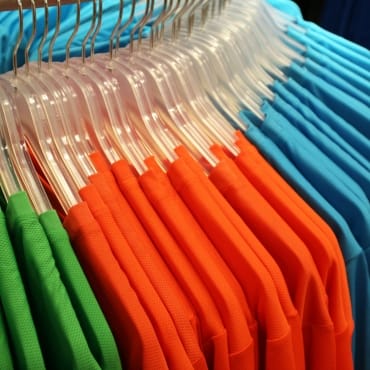 Colorful racks of polyester T-shirts in green, orange, and blue, hanging on clear plastic hangers.