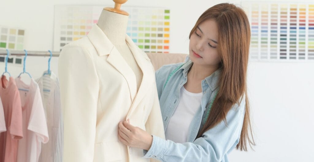 A woman examining a cream-colored blazer on a mannequin in a clothing store or design studio, with fabric swatches in the background.