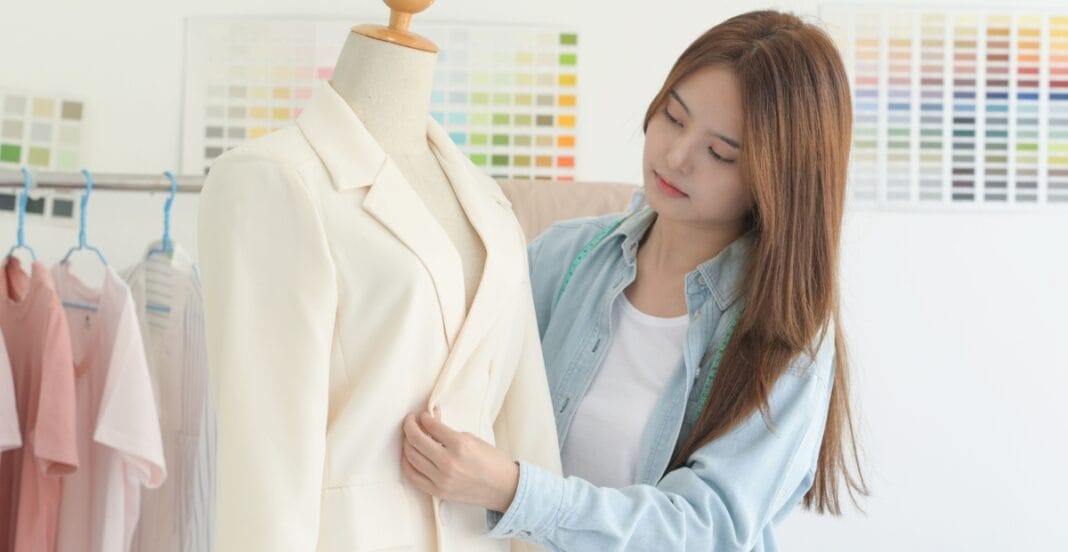 A woman examining a cream-colored blazer on a mannequin in a clothing store or design studio, with fabric swatches in the background.