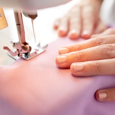 Close-up of a sewing machine stitching onto a piece of pink polyester fabric, with a person's hands guiding the fabric.