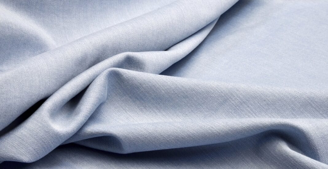 A close-up image of light blue stretch chambray fabric, softly draped with gentle folds and creases.