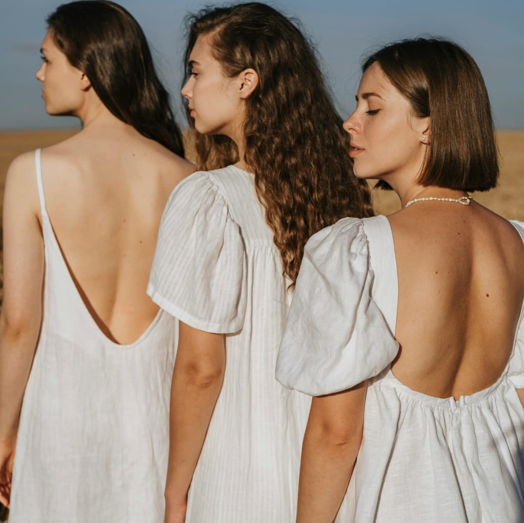 Three women in white gauze dresses standing outdoors on a sunny day, with their eyes closed or looking away, showcasing a breezy summery look.