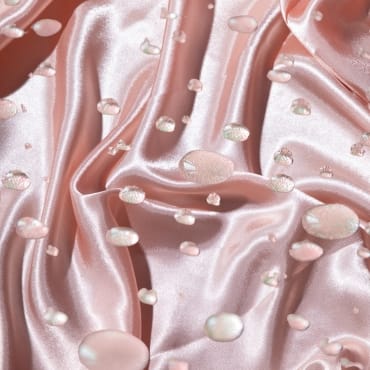 A close-up of soft, pink satin-like microfiber fabric that appears water-resistant, with a smooth, shiny surface and water droplets on top.
