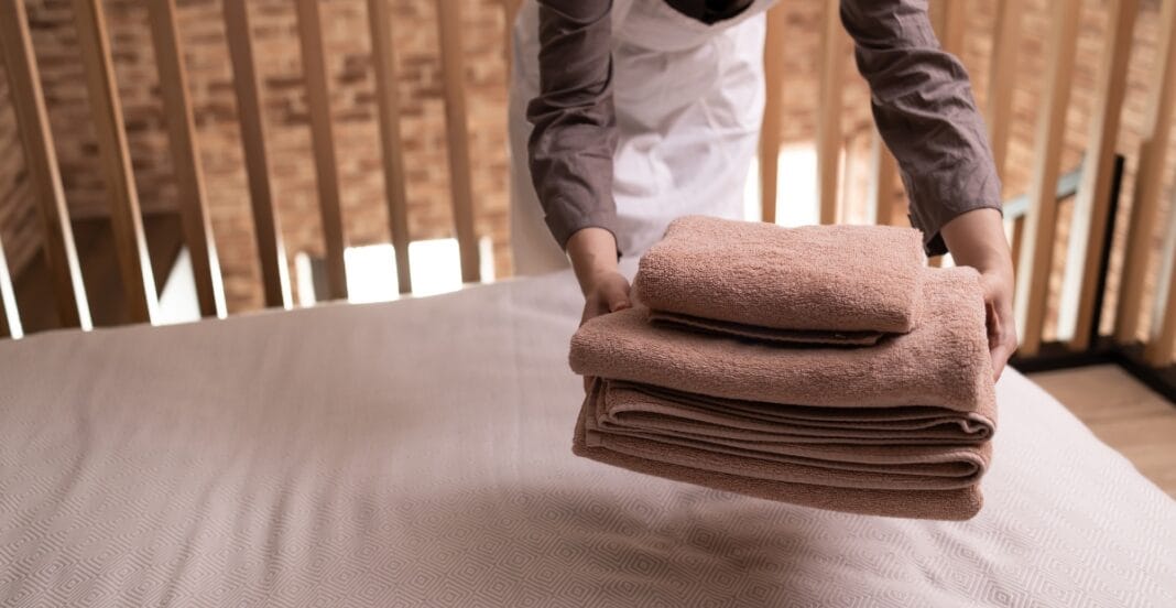 A person is folding a stack of beige towels on a bed, with a wooden slat wall in the background.