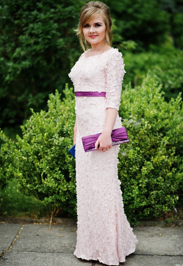 Elegant woman wearing a long, pale pink lace prom dress with a purple sash, holding a purple clutch bag and standing outdoors with green shrubbery in the background.