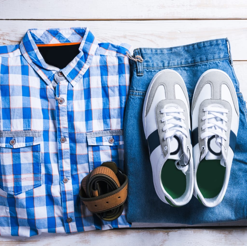 A pair of white sneakers, a brown belt, and a black watch are laid out alongside a blue and white checkered linen shirt on a wooden surface.