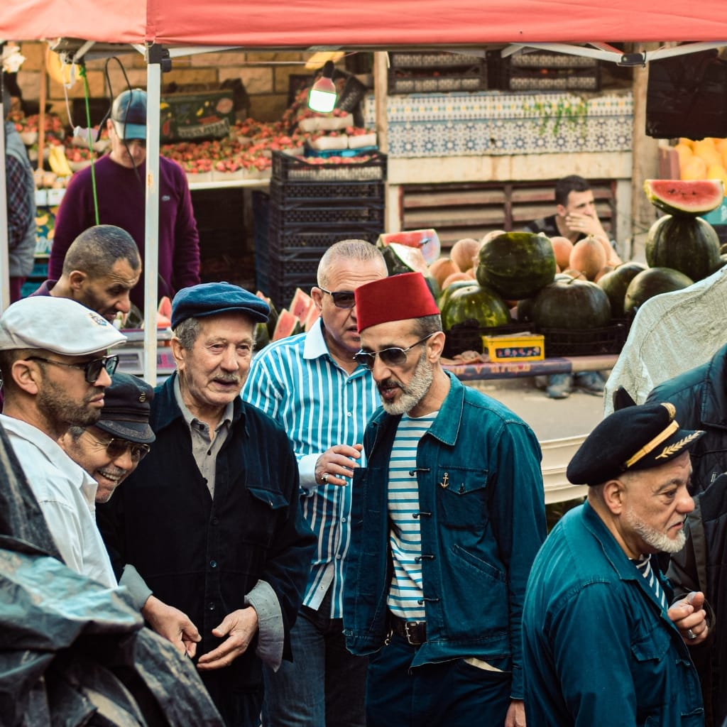 A group of men dressed in casual and nautical-inspired clothing, engaged in conversation at an outdoor market with watermelons and fruit stalls in the background.