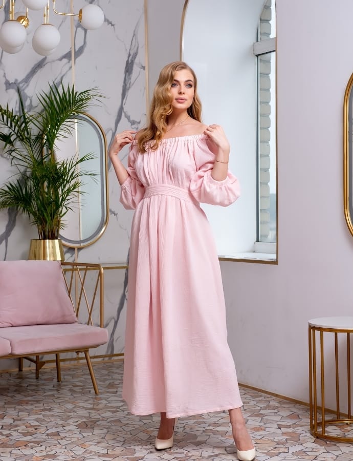 A woman wearing a light pink smock cotton dress standing in a modern, stylish room with marble walls, a mirror, a pink sofa, and gold accents.