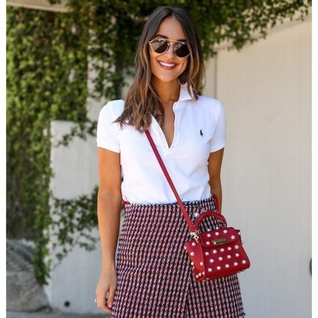 A woman smiling outdoors, wearing sunglasses, a white polo shirt, and a patterned skirt, carrying a red polka dot handbag.