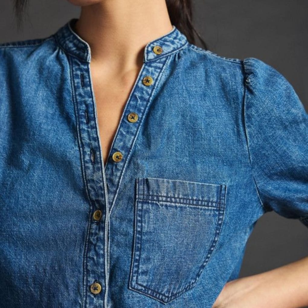 A close-up image of a woman wearing a denim chambray shirt with a stand-up collar and gold buttons. The shirt has a front pocket on the left side and slightly puffed sleeves, showing a casual yet stylish look.