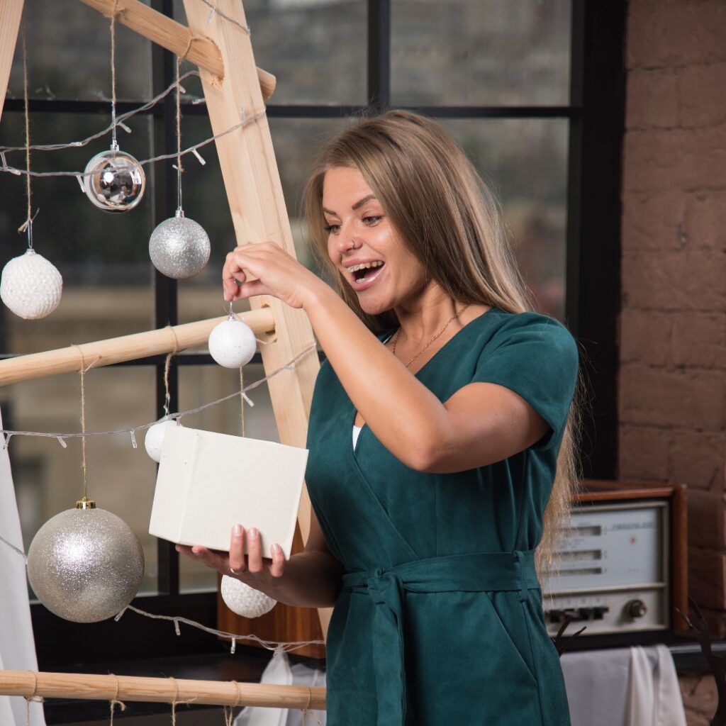 A woman with long brown hair wearing a teal dress is decorating a small white gift box with a Christmas ornament, standing next to a wooden frame decorated with hanging ornaments, in a cozy indoor setting with large windows.
