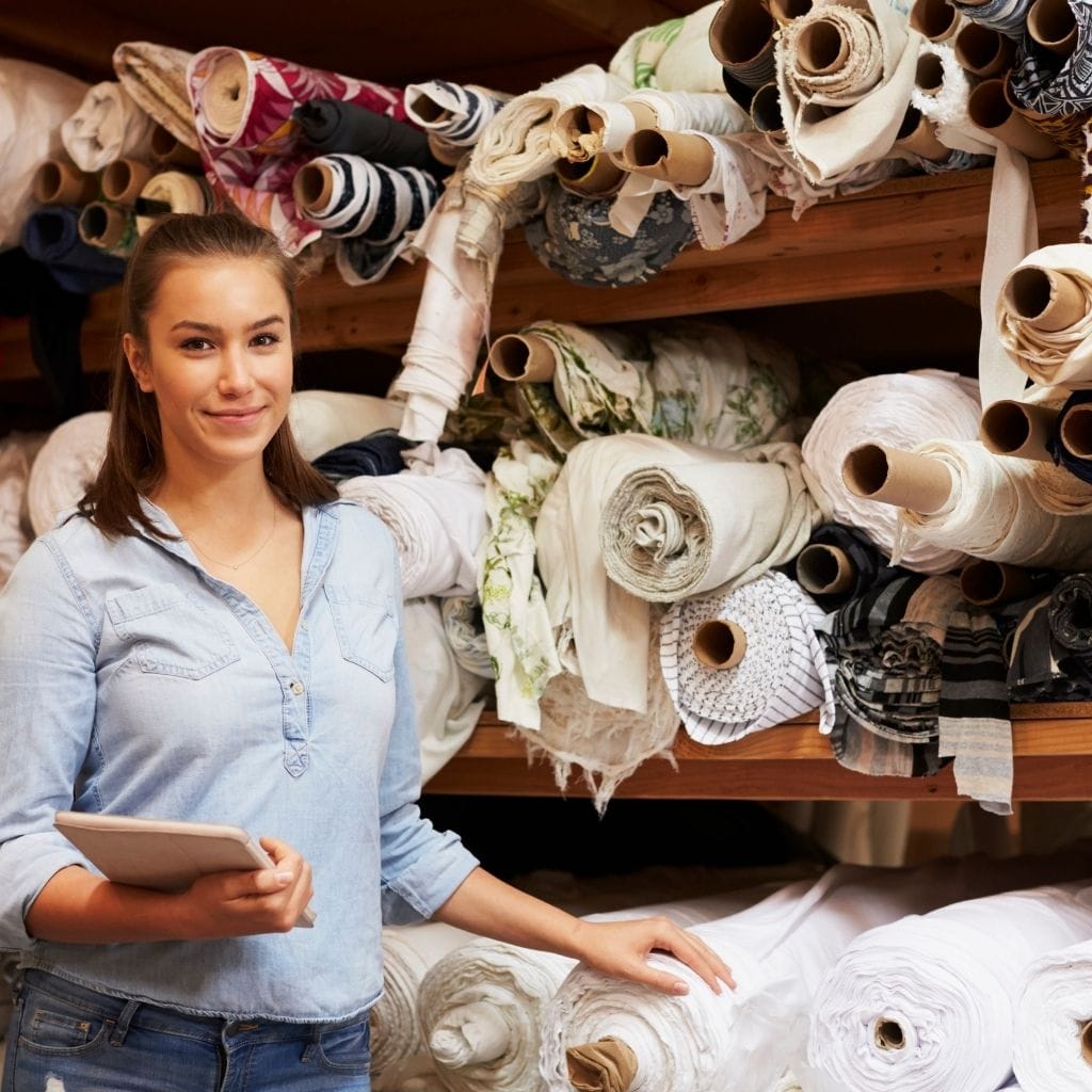 A young woman smiling at the camera while holding a notebook, standing in front of shelves stacked with rolls of fabric and textiles in various colors and patterns.