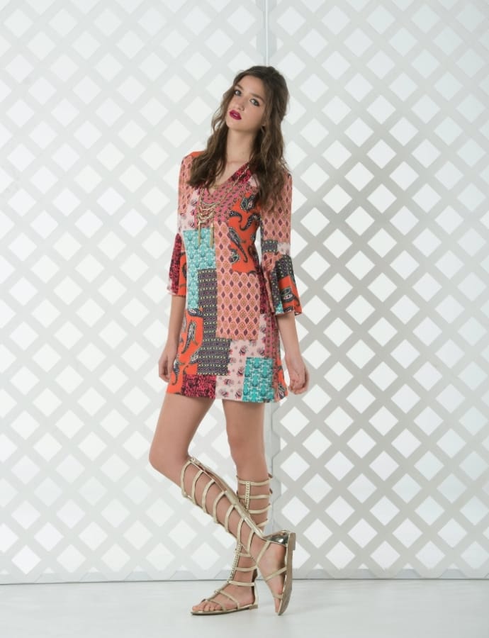 A young woman with long wavy hair and red lipstick, wearing a colorful patchwork dress with mixed patterns, paired with gladiator sandals, standing against a white geometric lattice background.