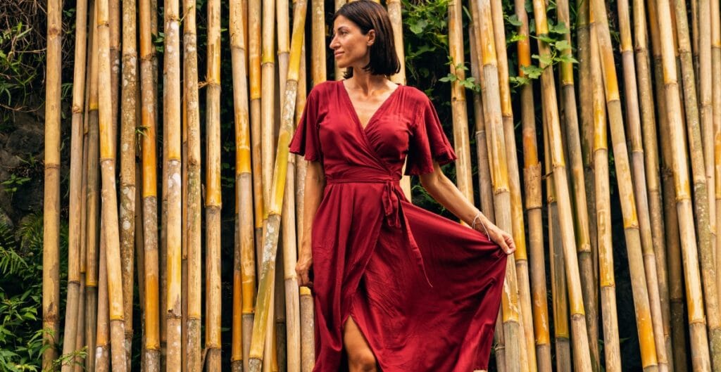 A woman in a red dress standing in front of a bamboo background, holding the side of her dress and looking to the right.